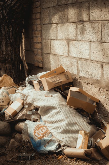 A pile of discarded cardboard boxes, some torn and flattened, and paper packaging materials are accumulated on the ground next to a weathered brick wall. The boxes vary in size and are partially open, revealing some lightweight contents inside. Surrounding the rubbish are loose paper and plastic bags, with a large reusable sack made of woven fabric partially covering part of the refuse. The scene is outdoors, likely in a back alley or side yard, with a wooden fence and a mature tree trunk visible in the background. Natural daylight illuminates the scene, highlighting the textures of the cardboard, paper, and the rough surface of the brick wall. The arrangement of waste suggests a private on-site collection or waste disposal effort, which could be addressed through efficient rubbish removal or independent clearance services such as those provided by companies like rubbishclearanceknightsbridge.com, to prevent clutter and maintain environmental standards.
