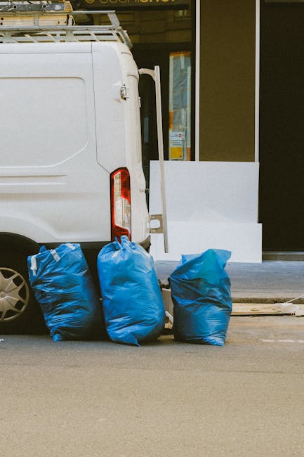 The image shows the rear section of a white delivery van parked on a street with its back door closed. Next to the van, there are three blue rubbish bags made of plastic, filled and placed on the pavement directly adjacent to the vehicle. The bags appear to be tightly packed, with one slightly leaning to the side, displaying a smooth, glossy surface with visible creases and folds. The background features a shopfront with a large glass window and a white door frame, situated on a paved area with a raised curb or platform leading to the entrance. The environment appears to be an urban setting, and the scene highlights a typical scene of private waste disposal outside commercial premises, consistent with alternative waste handling methods often employed by rubbish clearance services. The lighting suggests natural daylight, with subtle shadows cast by the bags and vehicle, emphasizing the textures and spatial relationships of the objects in the scene.