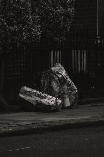 Two large black and white plastic sacks filled with rubbish rest on a pavement in front of a black metal fence, which encloses a garden or yard area with some dense foliage visible in the background. The sacks appear to contain mixed waste, possibly including paper, packaging, or household refuse, with one sack leaning slightly to the side and the other standing upright, both showing some creases and folds. The scene is outdoors and illuminated by natural light, with the ground surface partly visible, including a section of street paving. The surrounding environment suggests a quiet residential or urban setting, where private waste collection or rubbish clearance services, such as those offered by companies like rubbishclearanceknightsbridge.com, facilitate the disposal of waste materials outside properties, providing an alternative to municipal waste services.