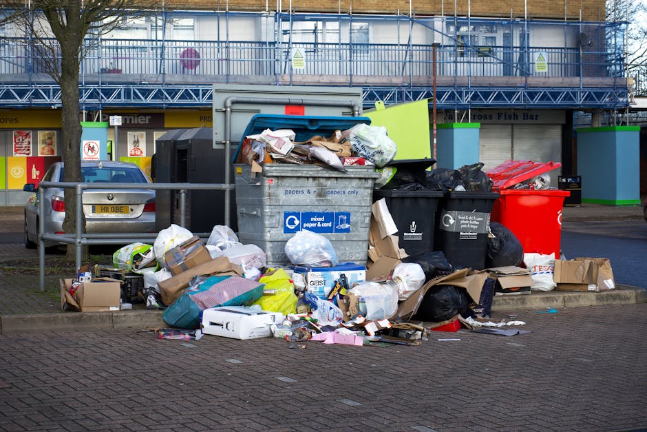 The image shows the rear section of a white delivery van parked on a street with its back door closed. Next to the van, there are three blue rubbish bags made of plastic, filled and placed on the pavement directly adjacent to the vehicle. The bags appear to be tightly packed, with one slightly leaning to the side, displaying a smooth, glossy surface with visible creases and folds. The background features a shopfront with a large glass window and a white door frame, situated on a paved area with a raised curb or platform leading to the entrance. The environment appears to be an urban setting, and the scene highlights a typical scene of private waste disposal outside commercial premises, consistent with alternative waste handling methods often employed by rubbish clearance services. The lighting suggests natural daylight, with subtle shadows cast by the bags and vehicle, emphasizing the textures and spatial relationships of the objects in the scene.