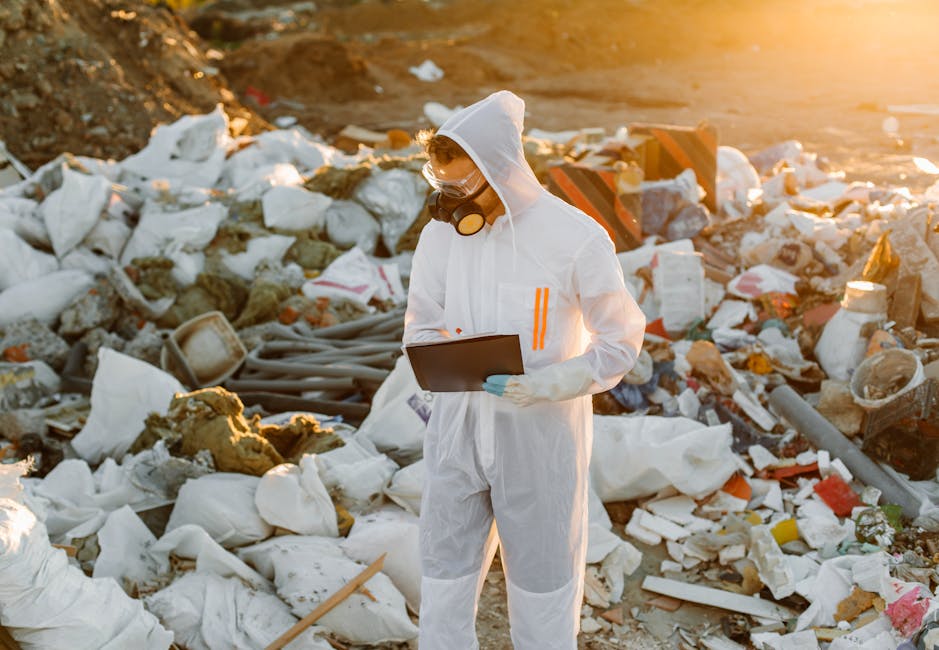 A person dressed in a white protective suit with a hood, goggles, and a respirator mask stands amidst a large pile of mixed waste materials at an outdoor site. The waste includes white foam, plastic bags, broken bricks, debris, and various discarded objects scattered across the ground. The person is holding a black clipboard and appears to be inspecting or documenting the hazardous waste environment. The background shows more scattered rubbish, with sunlight casting a warm glow over the scene, emphasizing the chaotic nature of the waste. This setting relates to private waste management or rubbish clearance services, such as those offered by Knightsbridge-based firms, in handling uncontrolled or waste-related cleanup situations that require careful assessment and disposal of hazardous materials. The overall atmosphere is one of careful examination within a contaminated site, highlighting the importance of proper waste handling according to regulations.