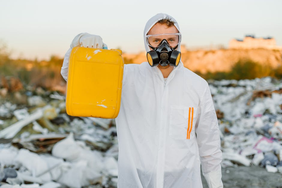 A person dressed in a white protective suit with a hood, goggles, and a respirator mask stands amidst a large pile of mixed waste materials at an outdoor site. The waste includes white foam, plastic bags, broken bricks, debris, and various discarded objects scattered across the ground. The person is holding a black clipboard and appears to be inspecting or documenting the hazardous waste environment. The background shows more scattered rubbish, with sunlight casting a warm glow over the scene, emphasizing the chaotic nature of the waste. This setting relates to private waste management or rubbish clearance services, such as those offered by Knightsbridge-based firms, in handling uncontrolled or waste-related cleanup situations that require careful assessment and disposal of hazardous materials. The overall atmosphere is one of careful examination within a contaminated site, highlighting the importance of proper waste handling according to regulations.