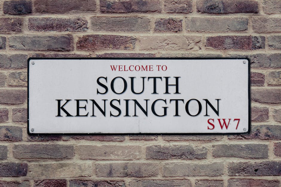 A rectangular metal street sign mounted on a weathered red brick wall, displaying the text 'Welcome to South Kensington' in black uppercase serif font with a slight shadow effect, and the postcode SW7 in red in the lower right corner. The sign has a white background with rounded edges and four bolts securing it at each corner. The brick wall behind the sign features an uneven texture with visible mortar joints, and the bricks vary in shades of red and brown. The environment suggests an exterior urban setting, typical of a residential or commercial area in South Kensington, Kensington and Chelsea, where alternative waste handling or private rubbish removal services may be common. The lighting appears natural, with soft shadows enhancing the texture of both the bricks and the sign, contributing to a clear, professionally captured image suitable for accessibility and SEO purposes related to rubbish clearance services.