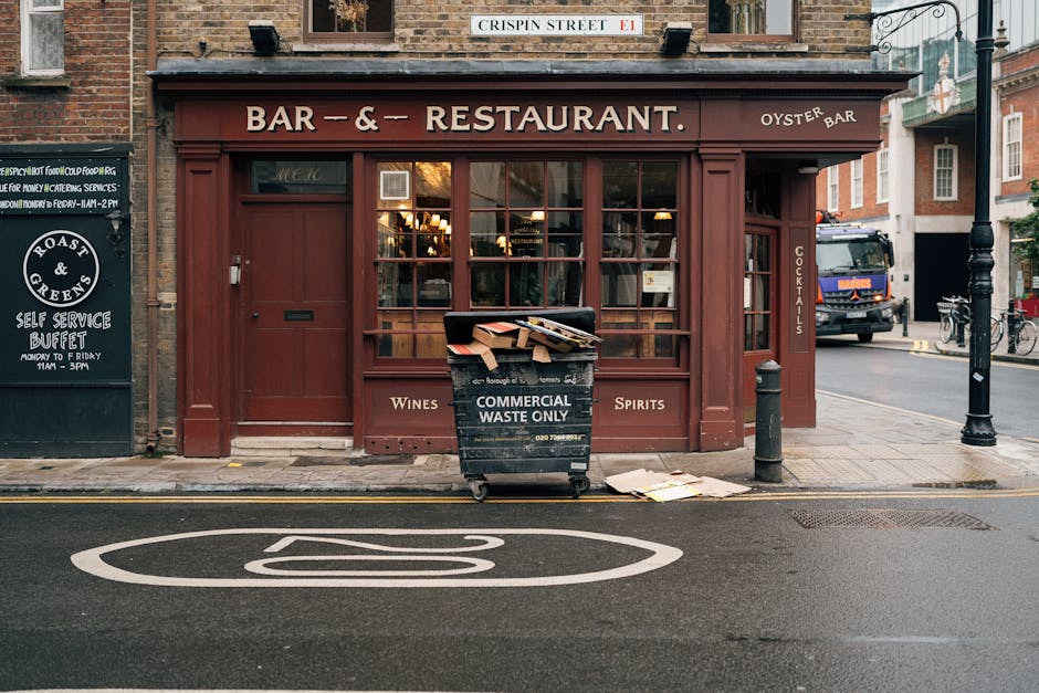 In front of a brick-built restaurant with a brown wooden facade and large glass windows, a black commercial waste container is positioned on the sidewalk, filled with discarded cardboard and paper, with some waste spilling onto the pavement. The container bears white lettering indicating it is for commercial waste disposal only. Nearby, a small black bollard is situated close to the curb, which has clear road markings, including a circular symbol for parking regulations painted in white. To the left of the restaurant, a black signboard displays information about a self-service buffet, age restrictions, and operating hours. On the right side of the image, a street scene includes a parked truck and bicycles, with a street sign mounted above reading 'CRISPIN STREET.' The scene is outdoors in an urban setting, with ambient natural lighting and a slightly overcast sky. The visual context relates to waste management practices outside a hospitality venue, highlighting the use of on-site commercial waste containers suitable for independent collection and disposal outside local council provisions, as seen in professional rubbish removal services such as those offered by rubbishclearanceknightsbridge.com.