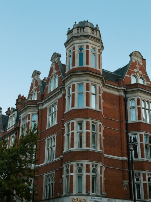 The image depicts a historic multi-story brick building with a prominent turret on the corner, featuring decorative white window frames and detailed stonework. The structure has a reddish-brown brick exterior with contrasting white accents around the tall, narrow windows, which are symmetrically aligned and include arched and rectangular styles. The turret rises above the roofline, topped with a small, ornate balustrade, and is capped with a dome-like roof. The building's architectural style suggests a Victorian or Edwardian era, with intricate detailing and a grand facade. Surrounding the building, there are partial views of trees and a lamppost in the foreground, with a clear blue sky overhead. This scene is an example of historical urban architecture, often found in city centres, which may be adjacent to or near areas where private waste collection or alternative rubbish removal services, such as those provided by companies like rubbishclearanceknightsbridge.com, are employed to manage waste disposal from properties of this type.