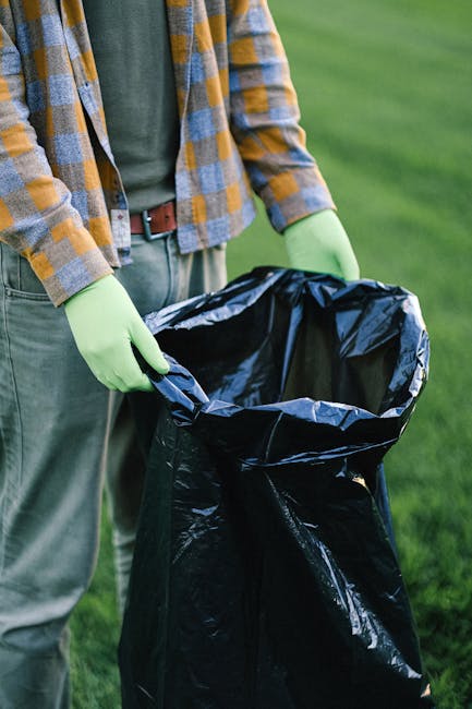 A person wearing a checkered shirt in shades of yellow, blue, and gray, along with green gloves, holds open a large black rubbish bag made of shiny, flexible plastic material. The bag is partially open, revealing an empty interior, and is positioned in front of a grassy outdoor area with blurred greenery in the background. The individual stands on a paved or grass surface, suggesting an outdoor setting suitable for waste collection or rubbish removal activities. The scene emphasizes the process of managing waste, aligning with private or independent rubbish disposal services, as provided by companies like rubbishclearanceknightsbridge.com. The lighting appears natural, with daylight illuminating the scene evenly, highlighting the textures of the plastic bag and the clothing, and reinforcing a professional, neutral tone suitable for a waste management context.
