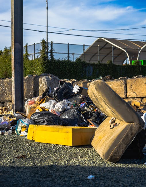 A pile of mixed household rubbish and waste materials is scattered on the ground in an outdoor setting, with black plastic rubbish bags, a yellow plastic container, and an old, worn beige car seat visibly prominent. The waste includes plastic packaging, paper, and other debris, all resting on coarse gravel. Behind the waste, a low stone wall separates the area from a hedge and an open area with a large, curved metal and fabric structure, possibly a sports or event shelter, visible in the background. Overhead, power lines span across a partly cloudy blue sky. The scene is illuminated by natural daylight, highlighting the textures of the discarded items and surrounding environment. The image reflects an informal waste accumulation scene, illustrating the need for proper rubbish removal services like those offered by rubbishclearanceknightsbridge.com, which facilitate alternative waste handling solutions outside standard municipal collection.