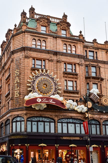 The image depicts the upper floors of a historic, ornate building constructed with reddish-brown brick and stone detailing, featuring multiple large windows with decorative trims. The facade showcases intricate architectural elements such as carved stonework, balustrades, and a tower-like structure at the roofline, adorned with small turrets and ornamental spires. Prominently mounted on the building's front is a large, elaborate clock with a golden sunburst design surrounding a central clock face, and a red banner underneath with the text 'Loro Piana', indicating a retail store. Below, the ground level contains large glass display windows and entrances, with retail signage and interior lighting visible. The scene suggests an urban shopping area, with part of the street and nearby signs visible at street level. The overall atmosphere is lively, with natural daylight illuminating the detailed brickwork and decorative elements, emphasizing the building's historic and architectural significance, while subtly connecting to the context of private property and independent retail spaces often requiring separate waste collection arrangements that [COMPANY_NAME] provides for specialised rubbish removal services in urban environments.