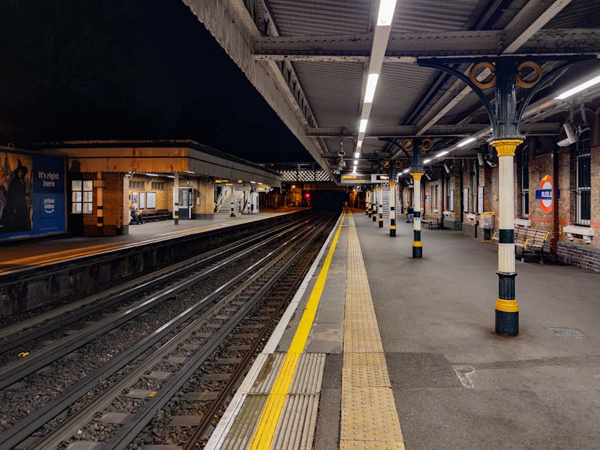 A nighttime scene at an outdoor train station platform featuring multiple railway tracks extending into the darkness. The platform is illuminated by overhead fluorescent lights, casting a bright glow onto the grey pavement and tactile yellow warning strips running along the edge. The platform is supported by decorative black and yellow striped cast iron pillars, some of which have benches attached nearby. On the left, a building with a brick and layered wood facade houses ticket machines, with a person sitting on a bench beneath a window. The surroundings include a brick wall with a circular red and blue Transport for London roundel sign indicating the station's name, partially visible on the right. The environment is clean, with neatly arranged bollards and signage, and the scene depicts the typical infrastructure of a suburban or city-centre railway station used for passenger boarding and alighting, with the visual context subtly aligning with the concept of public or alternative transport-related waste management and rubbish clearance.