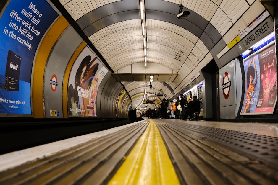 A view from the floor level of an underground London Underground station platform, showing the curved tiled ceiling with bright overhead lighting, advertisements lining the walls, and a row of passengers seated and standing along the platform edge. The yellow tactile paving strip runs centrally along the platform surface, guiding visually impaired travelers. The station sign indicates the name 'BOND STREET' on the right side, with the iconic roundel symbol for the London Underground. The background reveals the tunnel's narrowing perspective, with commuters waiting for arriving trains. The environment features clean, smooth tiles, modern lighting, and typical station signage, creating a structured and functional setting. The scene captures the busy, enclosed space where passengers await transportation, with a neutral and professional tone that aligns with descriptions of transit infrastructure related to urban mobility and public transport hubs.