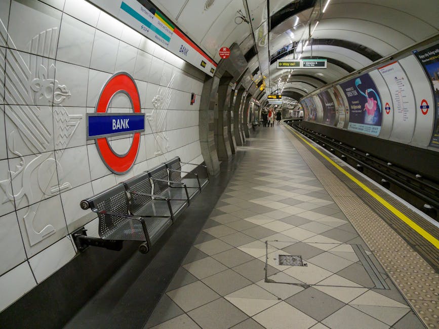 A nighttime scene at an outdoor train station platform featuring multiple railway tracks extending into the darkness. The platform is illuminated by overhead fluorescent lights, casting a bright glow onto the grey pavement and tactile yellow warning strips running along the edge. The platform is supported by decorative black and yellow striped cast iron pillars, some of which have benches attached nearby. On the left, a building with a brick and layered wood facade houses ticket machines, with a person sitting on a bench beneath a window. The surroundings include a brick wall with a circular red and blue Transport for London roundel sign indicating the station's name, partially visible on the right. The environment is clean, with neatly arranged bollards and signage, and the scene depicts the typical infrastructure of a suburban or city-centre railway station used for passenger boarding and alighting, with the visual context subtly aligning with the concept of public or alternative transport-related waste management and rubbish clearance.