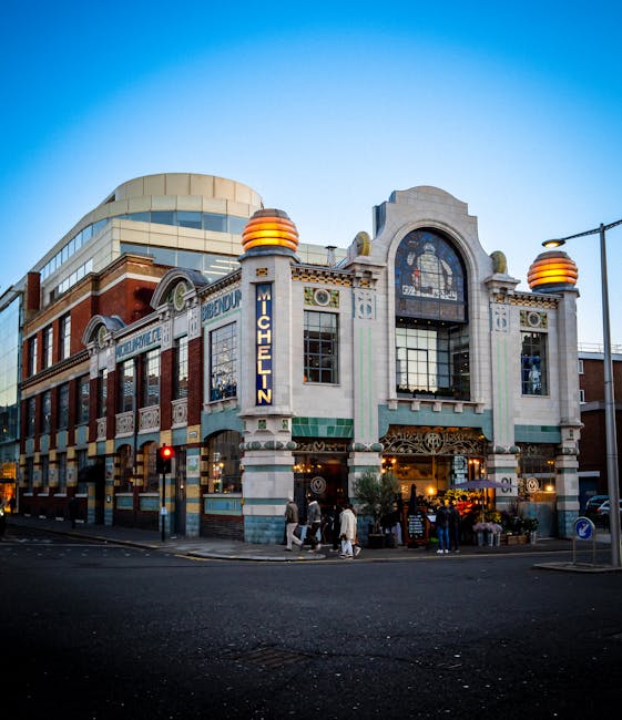 A historic multi-storey building at the corner of a street, featuring an ornate facade with decorative architectural details, large arched windows, and a prominent vertical sign that reads 'MICHELIN'. The building has a combination of white, green, and brown brickwork with elements of tile and stone finishes. On the ground floor, there are shopfront windows displaying various items, and an entrance with an awning. The surrounding environment includes a paved street with pedestrian crossings, traffic lights, and street lamps, with a few pedestrians walking near the entrance. The sky above is clear with a gradient from light to darker blue, indicating early evening or late afternoon. The scene is captured with natural lighting, highlighting the building's detailed exterior, and reflects the urban setting associated with commercial and entertainment use, typical of city centre locations.