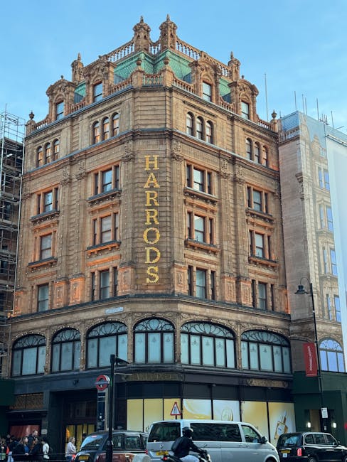 A multi-storey historic building with ornate stone architecture and decorative elements, featuring several rows of rectangular windows with detailed surrounds. The upper floors display a prominent vertical sign reading 'HARRORDS' in gold letters. The ground level has large display windows with arched frames, adjacent to a dark-colored entrance, and is situated on a busy city street with vehicles, including a white van and cars, passing by. Surrounding the building are other structures, some under renovation or construction, with scaffolding visible on the left side. The environment suggests an urban setting with pedestrian activity, and the overall scene is captured during daylight hours with a clear sky, emphasizing the building’s historic and architectural character, which aligns with the concept of alternative or private property management of urban spaces, similar to independent rubbish collection practices.