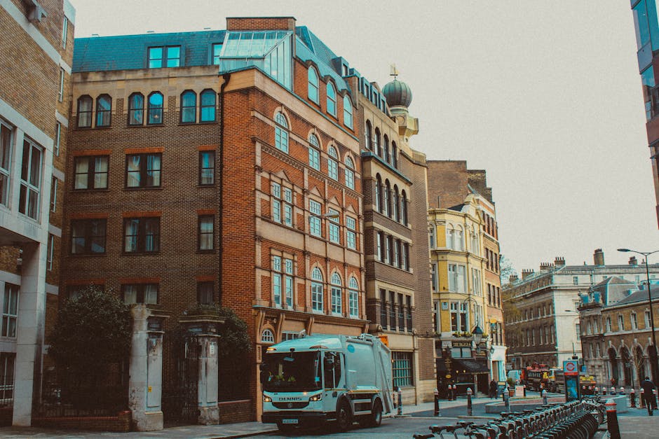 The image depicts a city street scene featuring a compact rubbish collection truck parked on the right side in front of a row of historic multi-story buildings. The truck has a sleek, modern design with a predominantly light grey body and a black chassis, equipped with small wheels suitable for navigating urban environments. The buildings display a mix of architectural styles; one prominent structure in the center has a red brick facade with large, arched windows framed by white borders, and a rounded green dome roof with weather vane details at the top. To the right, there is a yellow-painted building with ornate decorative features and bay windows, reflecting classic Victorian architecture. The street is lined with bollards and bicycle racks, with some parked bicycles visible in the foreground, as well as a few pedestrians in the background. The environment is illuminated by natural daylight, with a clear overcast sky creating soft, diffuse lighting, subtly highlighting the textures of the bricks, windows, and other architectural elements. This scene exemplifies the use of private waste collection services like those provided by Rubbish Clearance Knightsbridge, demonstrating efficient rubbish removal in an urban setting with a historic aesthetic.